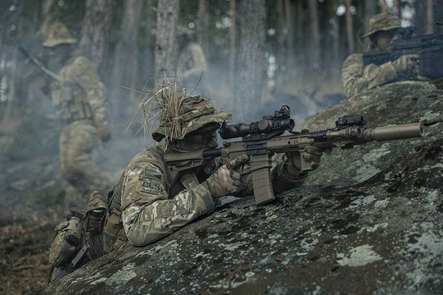 Rifleman with a scoped carbine on a rock while the team moves through the forest