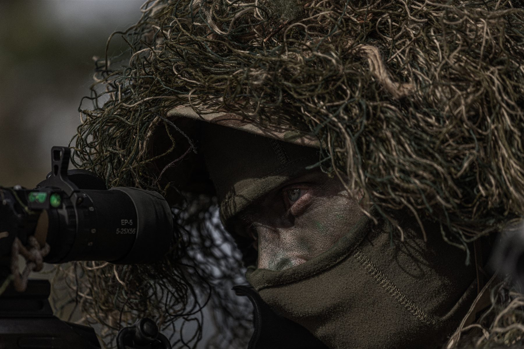 Sniper in a ghillie hood behind a night-vision scope